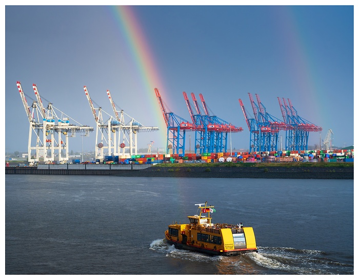 Blick auf ein Schiff vor mehreren Kränen am Hamburger Hafen mit einem großen Regenbogen