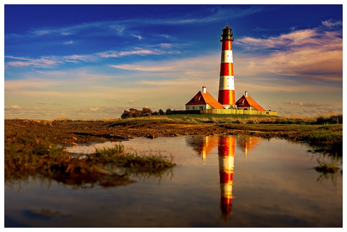 Blick auf den schicken Leuchtturm von Westerheversand an der Nordsee mit Spiegelung in einer Wasserpfütze bei Sonnenuntergang